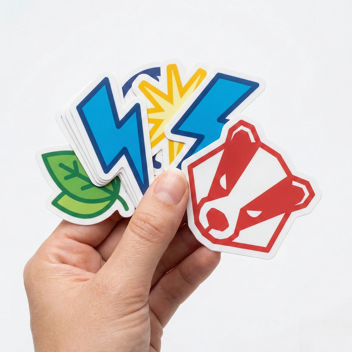 A hand holding several icon-style stickers, including blue and yellow lightning bolts, a green leaf, and a red animal logo, on a white background, Okaystickers
