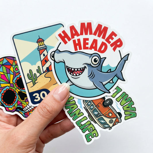 Hand holding a stack of glossy laminated cartoon stickers including a hammerhead shark, lighthouse, skull, and van designs
