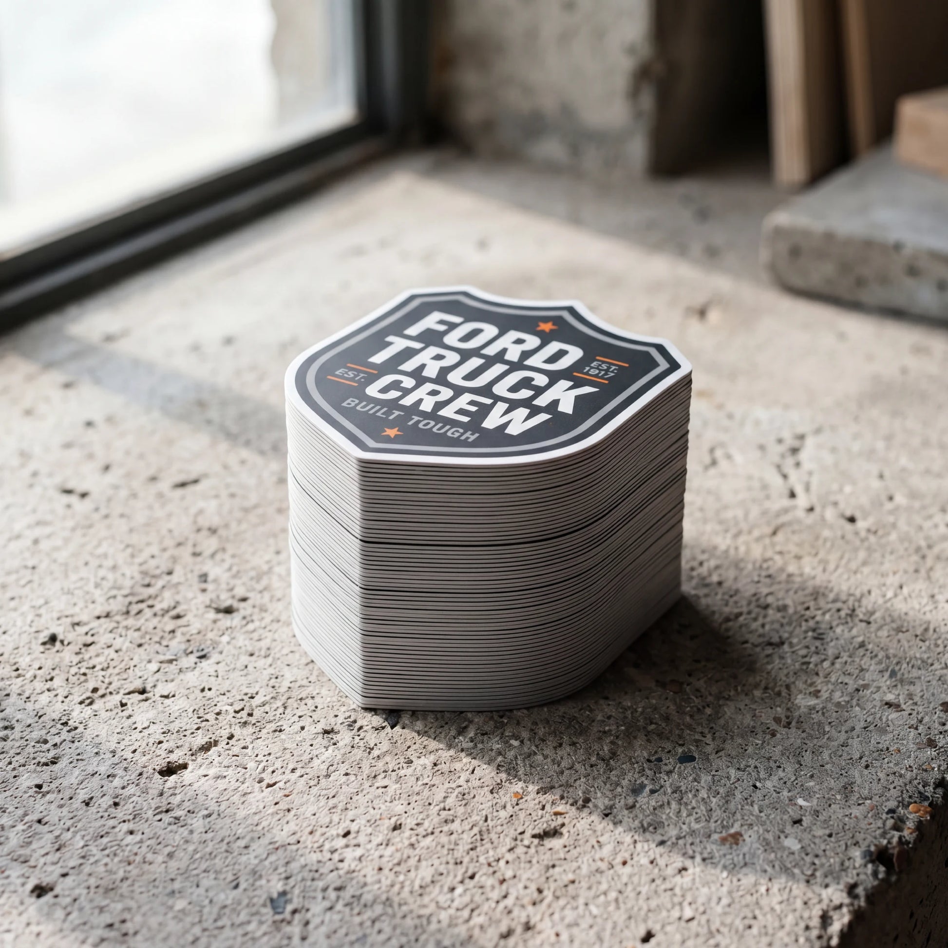 A thick stack of custom die-cut vinyl stickers in a shield shape, featuring "FORD TRUCK CREW BUILT TOUGH" branding, displayed on a concrete floor near a window.
