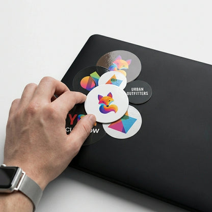 A focused shot of a black matte laptop lid covered in a colorful collage of personalized round vinyl labels and stickers, including a stylized fox logo and geometric designs.